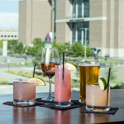 The image shows five different drinks, including cocktails, a beer, and a wine glass, placed on a table with a large brick building in the background.