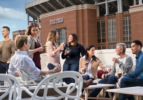 A group of people is gathered outdoors near a building, talking and enjoying drinks at a social gathering.