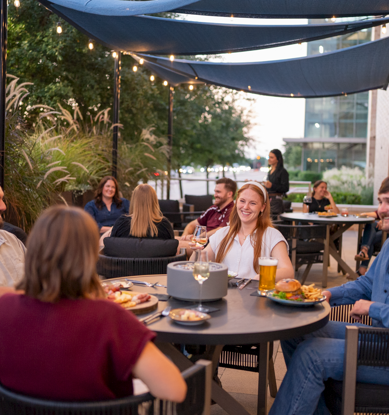 Friends dine and chat on a patio under a canopy with string lights and beers, creating a warm evening gathering.