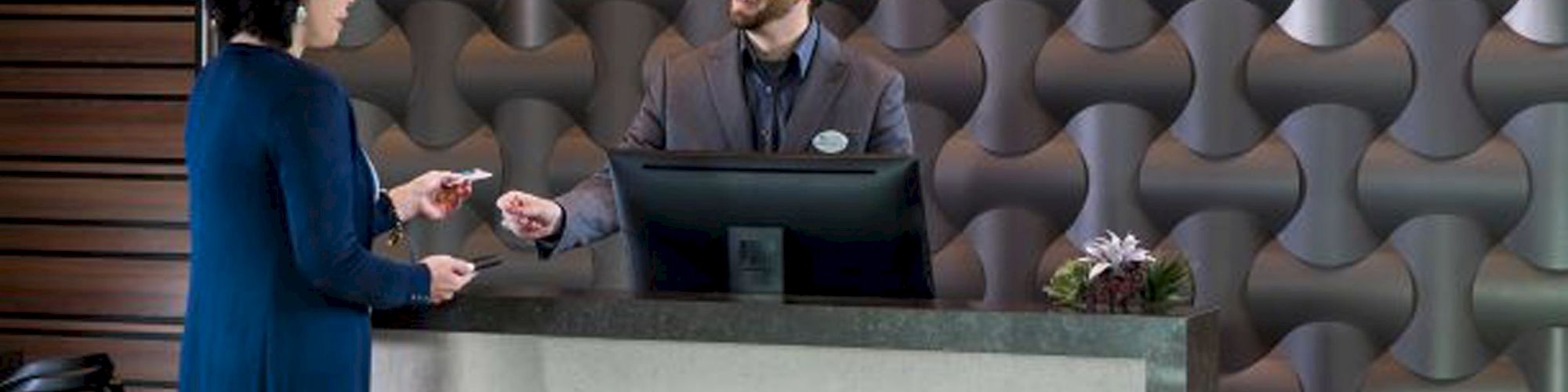 A woman hands a card to a receptionist at a counter with "ATM" engraved on the front, against a modern geometric patterned wall.