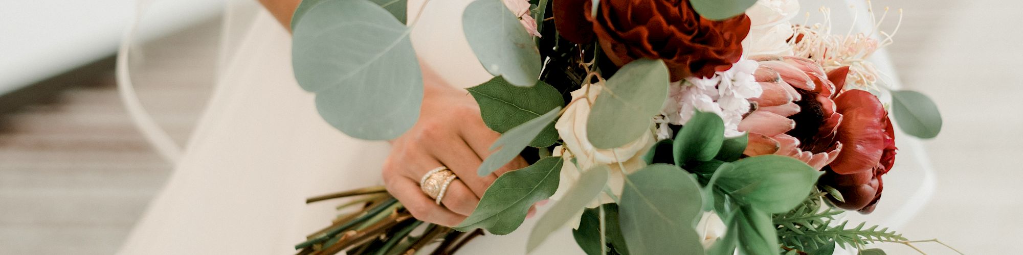 A bride in a white wedding dress holds a bouquet with green leaves and red flowers, standing in a well-lit indoor hallway.