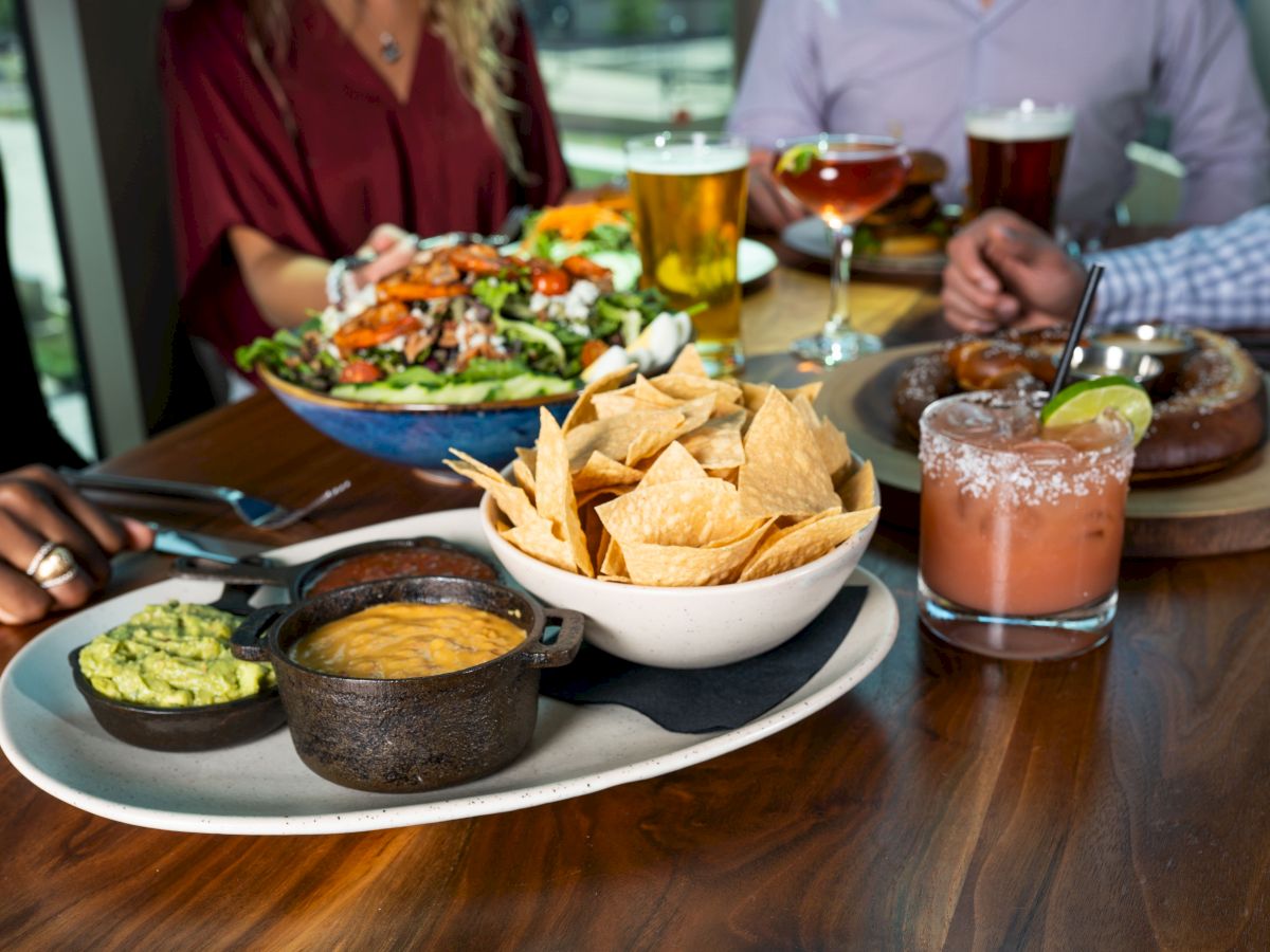 A table of shared Mexican-style food: tacos, salad, tortilla chips with dips, and drinks with glasses of beer.