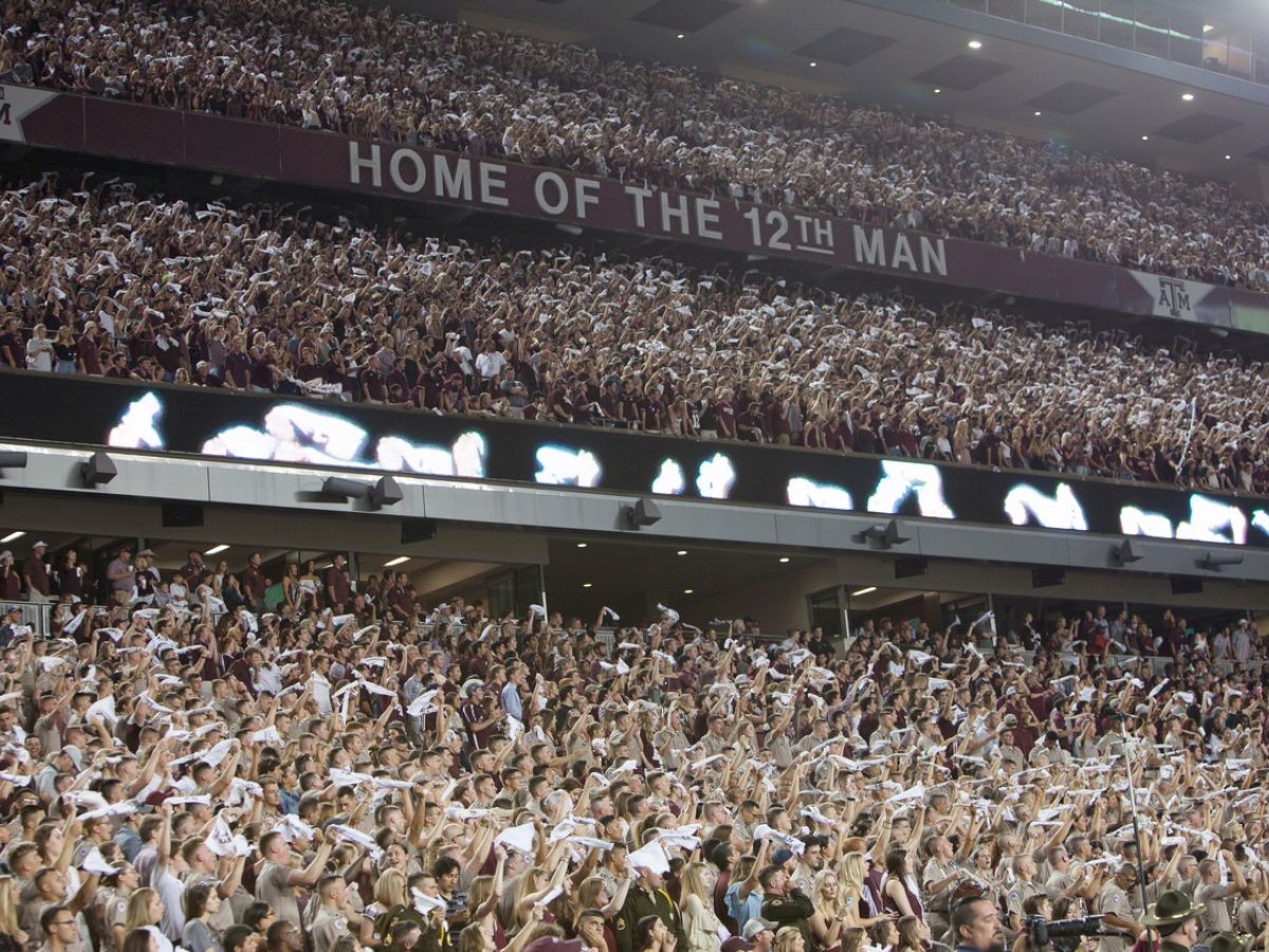 A large crowd at a stadium, with a banner reading "Home of the 12th Man." Fans are waving white towels in the air excitingly.