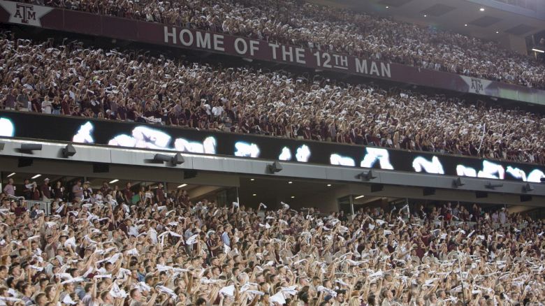 A large crowd at a stadium, with a banner reading "Home of the 12th Man." Fans are waving white towels in the air excitingly.