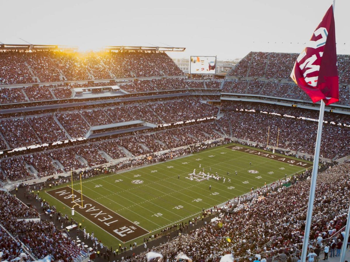 A large football stadium is packed with fans and flags waving, with a game underway on the field and the sun setting in the background ending the sentence.