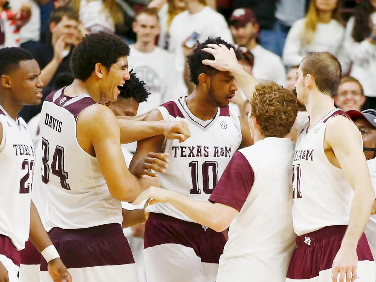 A group of Texas A&M basketball players are celebrating a victory on the court, with one player, number 10, in the center.