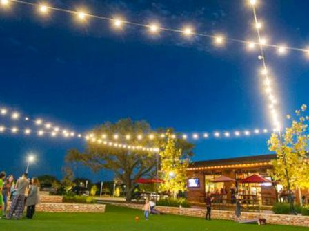 The image shows an outdoor area at night with string lights overhead, people gathered on a grassy space, and a building with a sign that says "Hopdoddy."