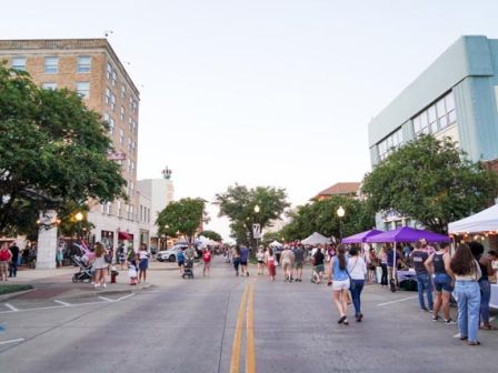 People are walking along a pedestrian street with vendor tents, buildings, and trees on either side during a street fair or market event.
