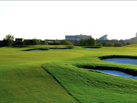 A well-maintained golf course with bunkers and undulating fairways, with a stadium structure visible in the background under a clear sky.