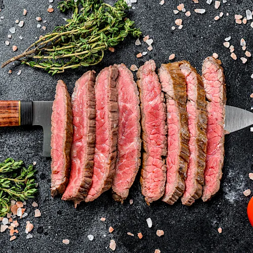 Sliced seasoned beef steak with herbs, salt, and pepper sits on a dark board beside a chef’s knife and cherry tomatoes.
