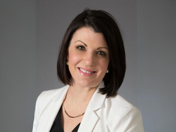 A person with shoulder-length dark hair, wearing a white blazer over a black top, smiles at the camera against a plain background.