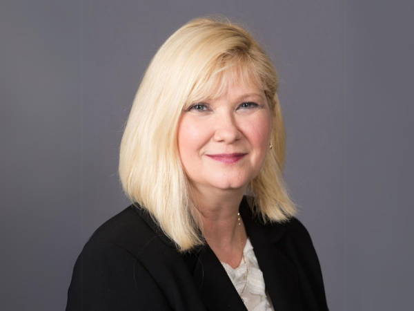 The image shows a woman with blonde hair, wearing a black blazer and a white blouse, smiling in front of a plain background.