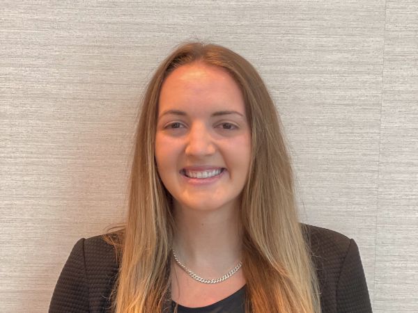A person with long hair, wearing a dark blazer and shirt, stands against a light-colored wall, smiling at the camera.