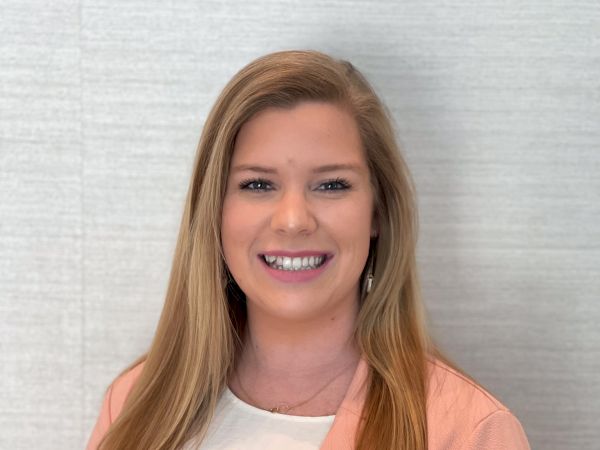 A smiling woman with long blond hair is wearing a light pink blazer over a white top, standing against a light-colored background.