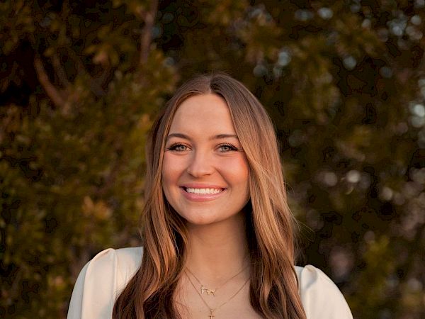 A person with long hair is smiling, standing outdoors with blurred green foliage in the background.