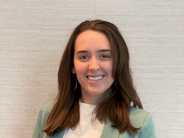 A person is standing in front of a plain background, wearing a light green blazer and a white shirt, with a friendly smile and dark brown hair.