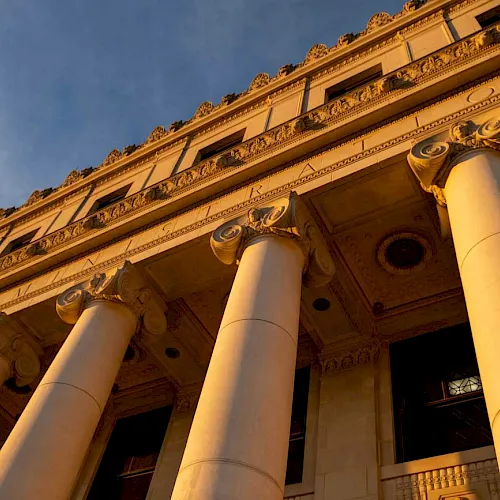 A grand neoclassical building fa&ccedil;ade with ornate gold trim and tall white columns under a blue sky.