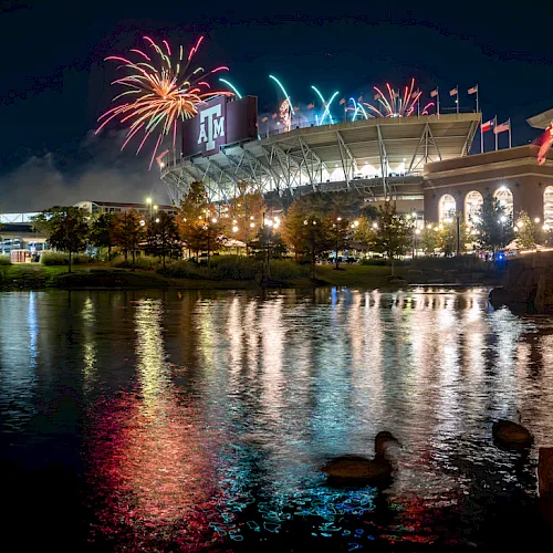 Fireworks burst over a lit stadium and flags, reflected in a calm lake at night.