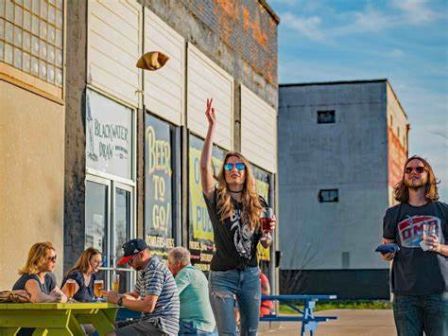 People are at an outdoor gathering, with a woman tossing a football and others seated at tables. A brewery or pub is visible in the background.