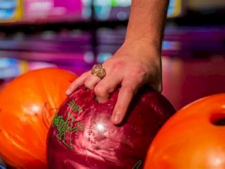 A hand is gripping a maroon bowling ball with a ring on the middle finger; two orange bowling balls are nearby at a bowling alley.