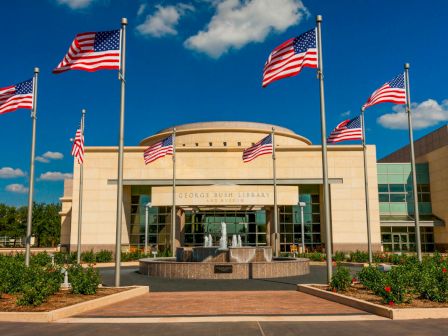 A stately building is seen with six American flags and a fountain in front, and the facade reads "George Bush Library." The sky is clear and blue.