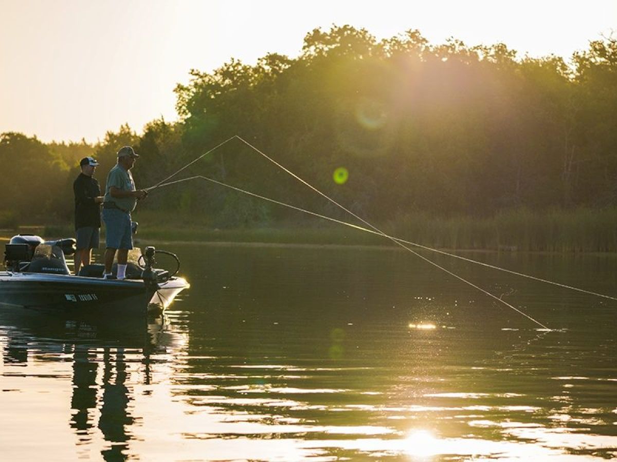 Two people are fishing on a boat in a calm lake during sunset, surrounded by trees in the background. The water glistens in the soft sunlight.