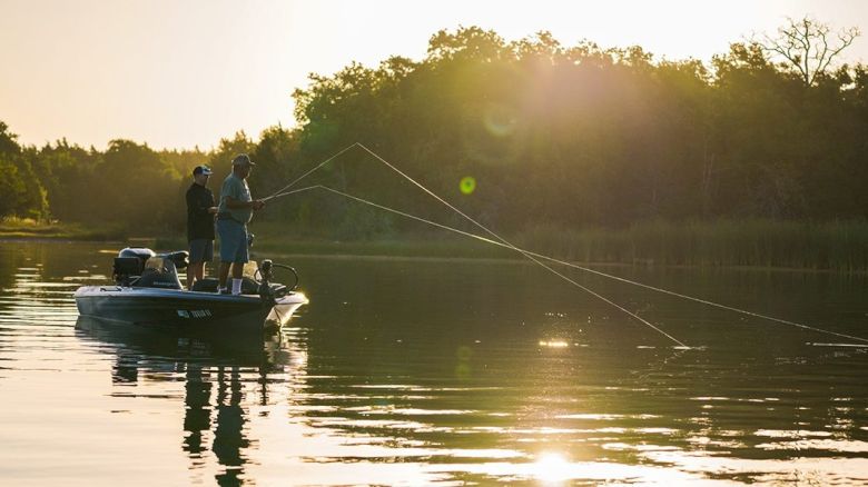 Two people are fishing on a boat in a calm lake during sunset, surrounded by trees in the background. The water glistens in the soft sunlight.