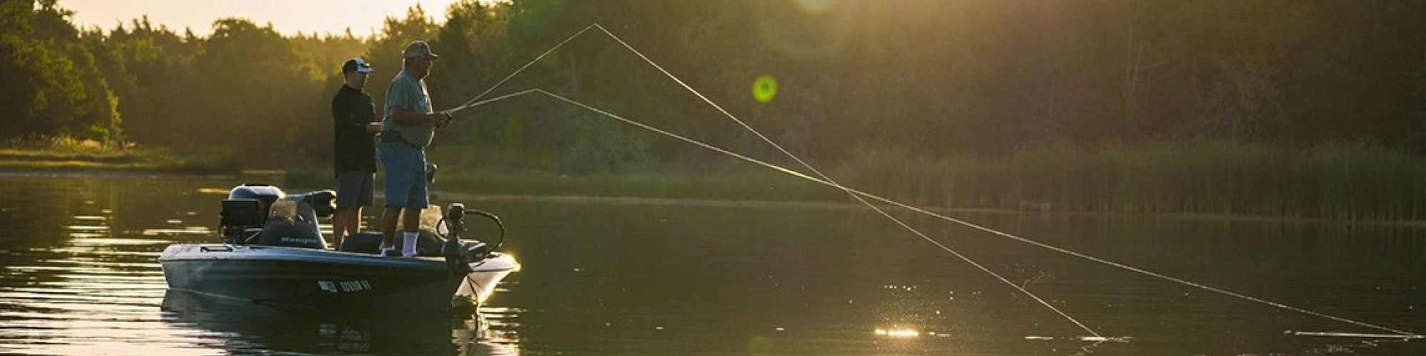 Two people are fishing from a boat on a calm lake during sunset, with trees in the background and reflections on the water.