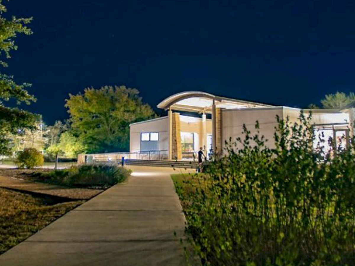 A modern building with a curved roof is illuminated at night, surrounded by greenery and trees, with a walkway leading up to it.