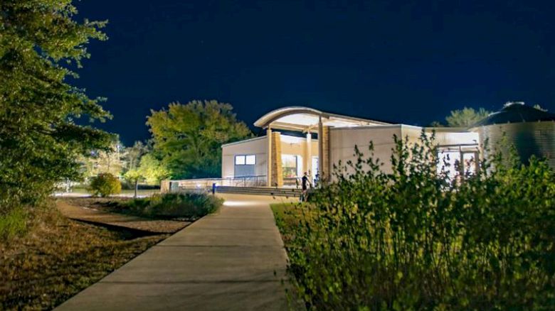 A modern building with a curved roof is illuminated at night, surrounded by greenery and trees, with a walkway leading up to it.