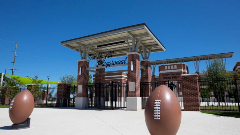 The image shows the entrance to a sports playground named "Crown Field." There are two large football sculptures flanking the path.