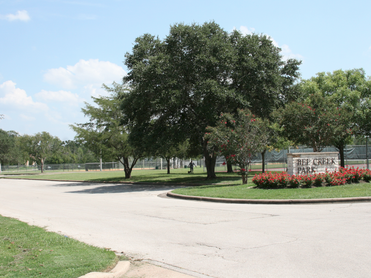 The image shows a park entrance with a sign reading "Deep Creek Park," surrounded by trees, a grassy area, and a tennis court in the background.