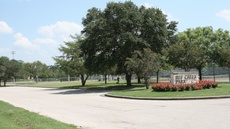 The image shows a park entrance with a sign reading "Deep Creek Park," surrounded by trees, a grassy area, and a tennis court in the background.