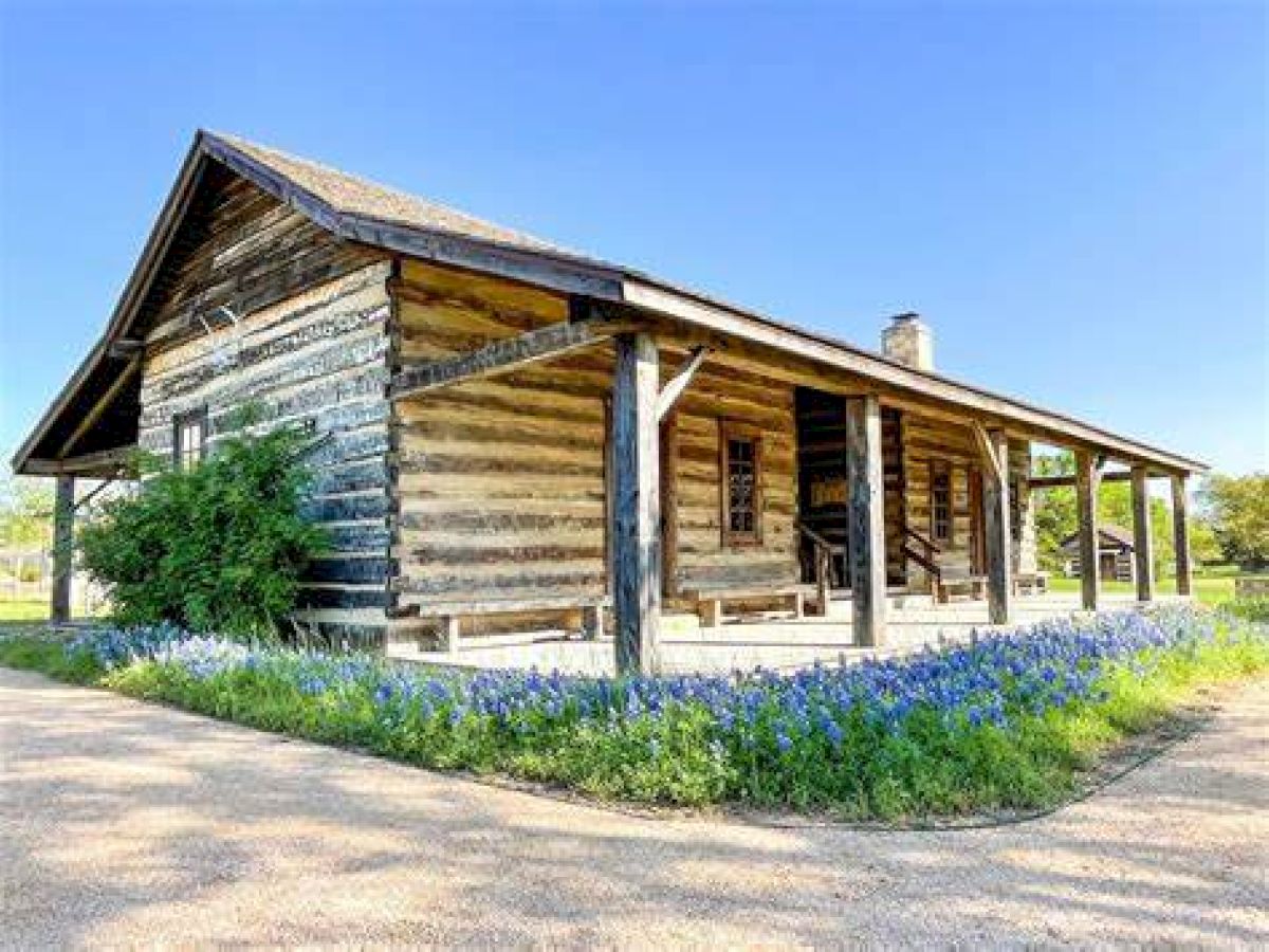 The image shows a rustic wooden cabin with a covered front porch and surrounding greenery, set against a clear blue sky, and flowerbeds in front.
