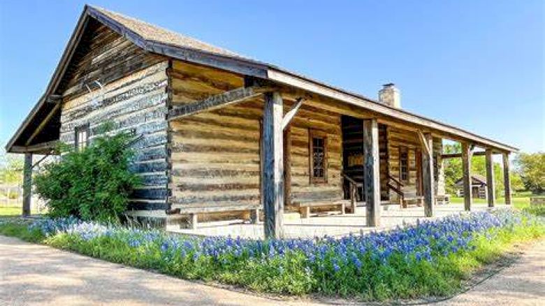 The image shows a rustic wooden cabin with a covered front porch and surrounding greenery, set against a clear blue sky, and flowerbeds in front.