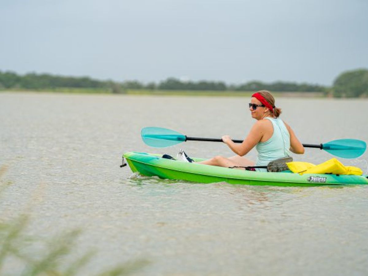 A person wearing a red headband and sunglasses is kayaking on a calm body of water with trees in the background, holding a double-bladed paddle.