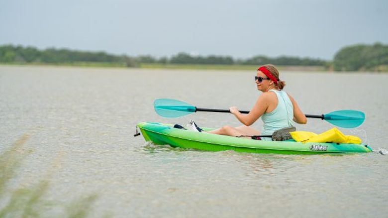 A person wearing a red headband and sunglasses is kayaking on a calm body of water with trees in the background, holding a double-bladed paddle.