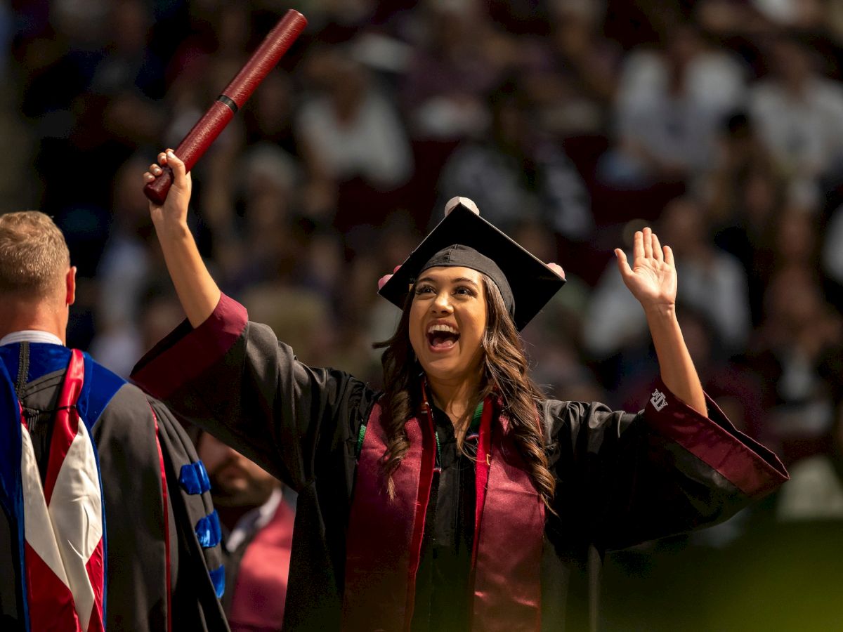 A person in graduation attire is celebrating with arms raised, holding a diploma, while another in academic regalia stands in the foreground.