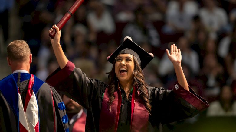 A person in graduation attire is celebrating with arms raised, holding a diploma, while another in academic regalia stands in the foreground.
