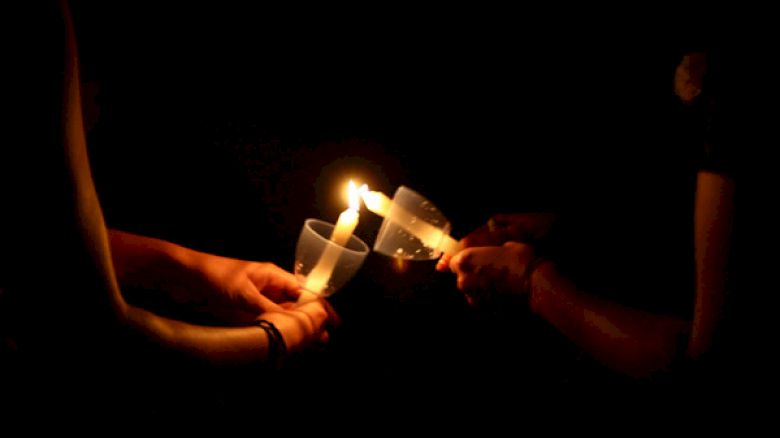 Three hands are holding candles with plastic covers in the dark, sharing the flame from one candle to another.