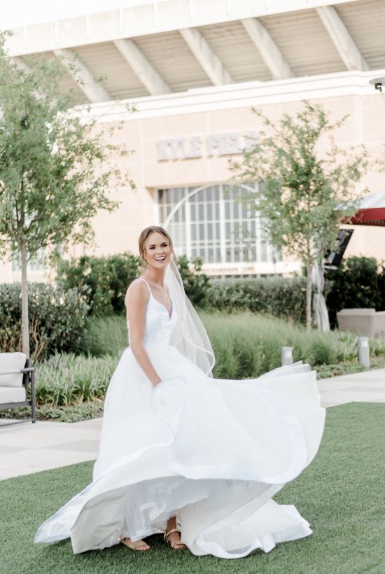 A woman in a white wedding dress is standing outdoors in front of a building, with trees and greenery in the background.