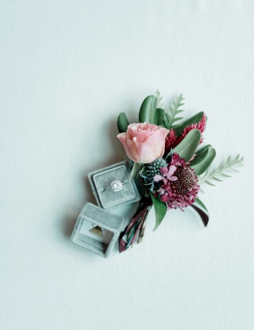 A diamond ring in an open box is next to a small bouquet of flowers with pink roses and greenery on a light background.