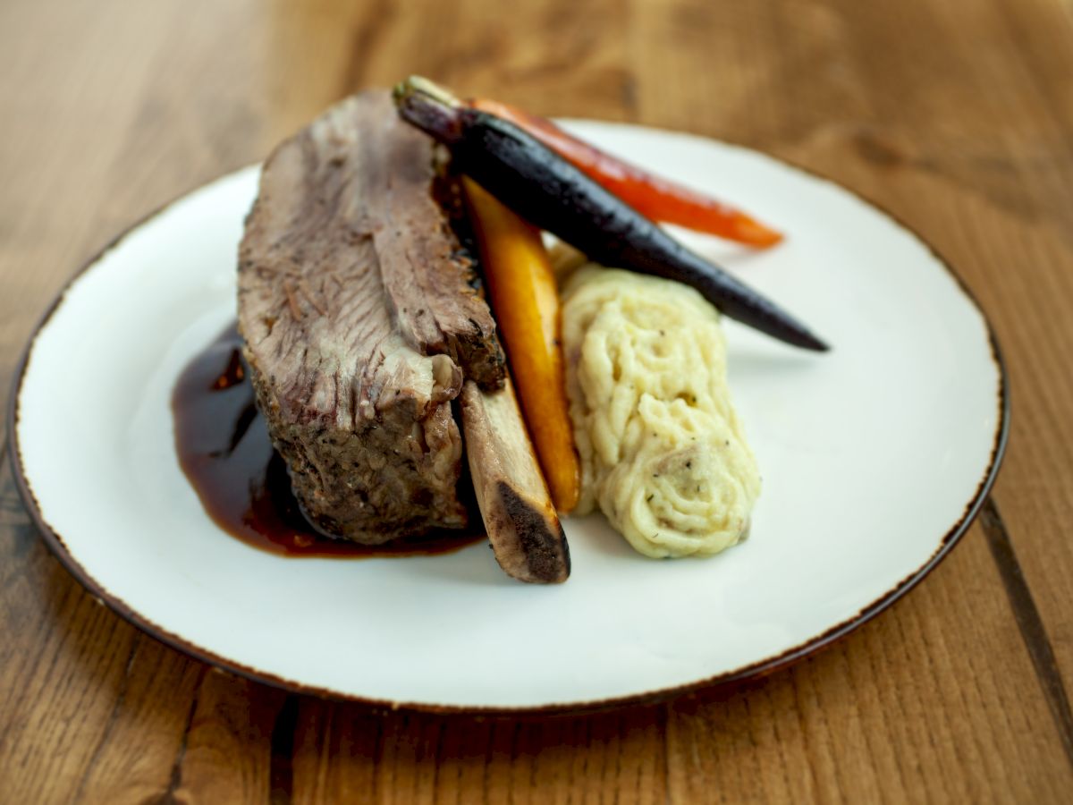 A plate with a rib, mashed potatoes, and roasted vegetables is set on a wooden table.