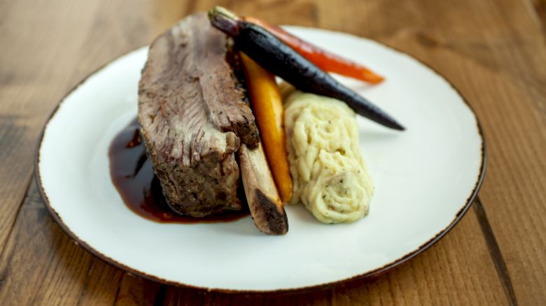 A plate with a rib, mashed potatoes, and roasted vegetables is set on a wooden table.