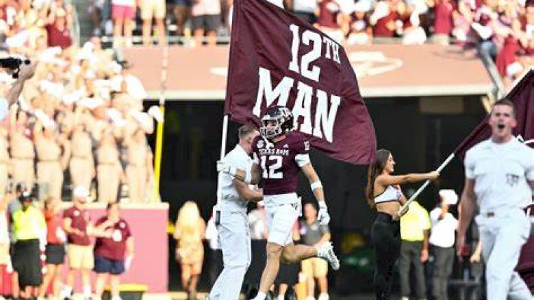 People on a football field holding flags that read "12th MAN," with a crowd in the background and someone cheering on the right.