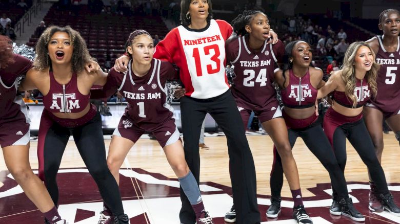 A group of women wearing Texas A&M outfits and one in a "NINETEEN 13" shirt are standing together on a basketball court, looking enthusiastic.