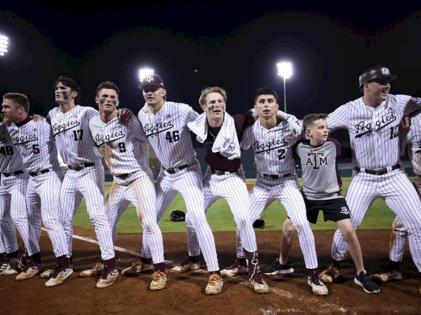 A baseball team in matching uniforms is lined up with arms interlinked, celebrating on the field under stadium lights.