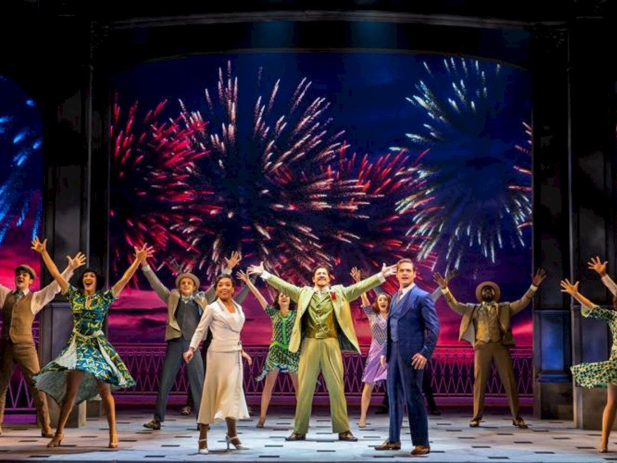 A stage performance with actors in colorful costumes, arms raised, in front of a backdrop displaying fireworks, creating an energetic scene.