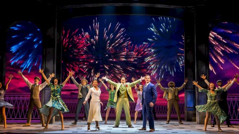 A stage performance with actors in colorful costumes, arms raised, in front of a backdrop displaying fireworks, creating an energetic scene.