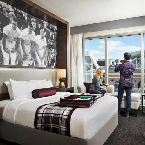 A hotel room with a bed, sports-themed decor, large window, two people looking outside, and various modern furnishings.